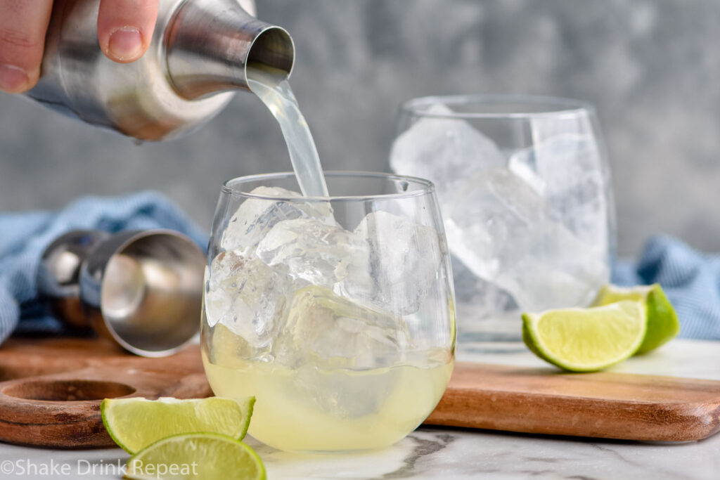 Man's hand pouring cocktail shaker of Canchanchara ingredients into a glass of ice surrounded by fresh lime wedges