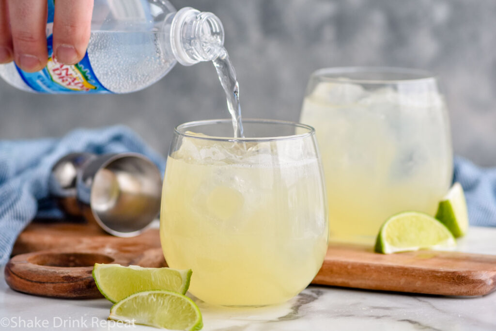 man's hand pouring club soda into glass of Canchanchara recipe with ice, surrounded by cocktail jigger and fresh lime wedges