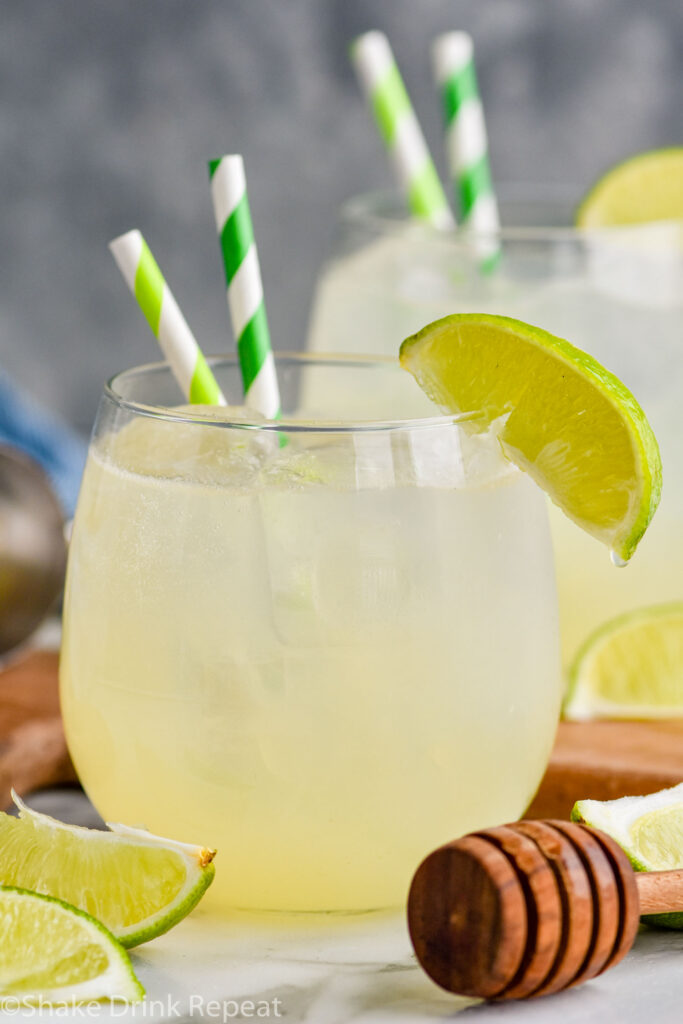 two glasses of Canchanchara recipe with ice, straws, and lime wedge with honey dipper laying in front of glass