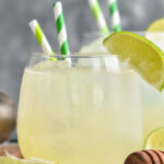 Glass of Canchanchara recipe with ice, straws, garnished with a lime surrounded by fresh limes and a honey dipper