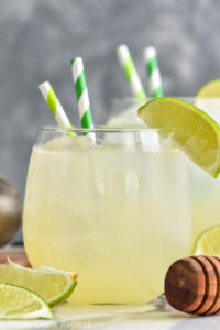 Glass of Canchanchara recipe with ice, straws, garnished with a lime surrounded by fresh limes and a honey dipper