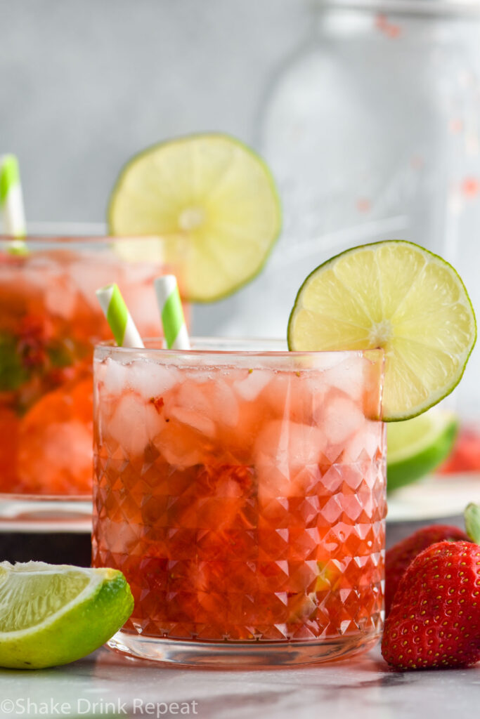 two glasses of Strawberry Caipiroska with crushed ice, a lime garnish and two straws surrounded by fresh strawberries and a lime wedge