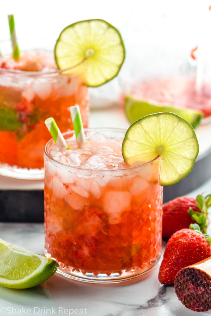 two glasses of Strawberry Caipiroska with crushed ice, two straws, and a lime slice surrounded by a cocktail muddler and fresh strawberries