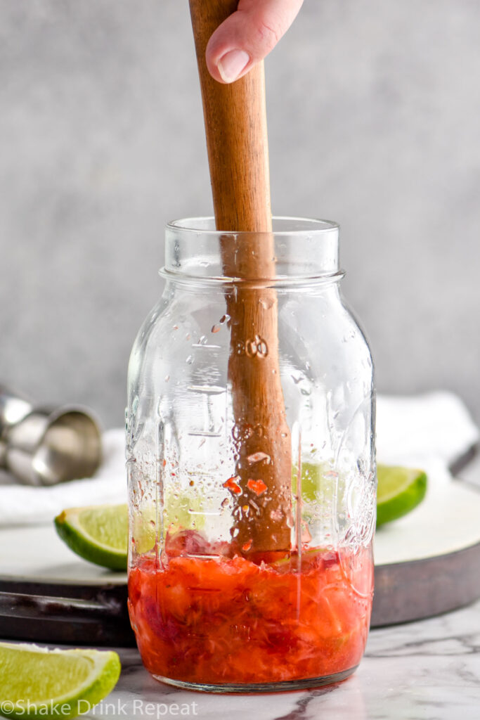 man's hand using cocktail muddler to mash strawberries, limes, and sugar in a mason jar to make a Strawberry Caipiroska recipe surrounded by fresh lime wedges