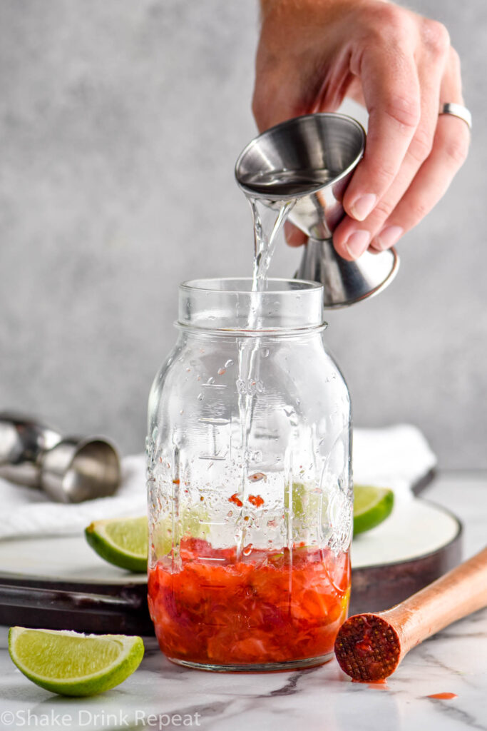 man's hand pouring cocktail jigger of Vodka into a mason jar of muddled strawberries and Strawberry Caipiroska ingredients surrounded by lime wedges and a cocktail muddler