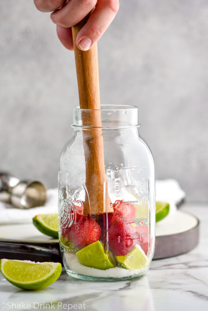 man's hand using a wooden cocktail muddler to mash fresh strawberries, limes, and sugar to make a Strawberry Caipiroska recipe