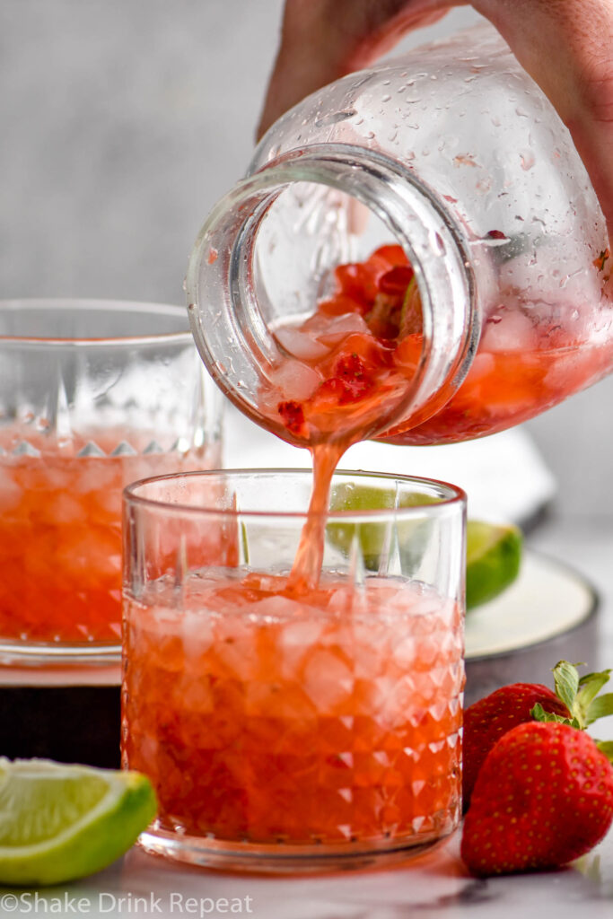 man's hand pouring mason jar of muddled strawberries into a glass of Strawberry Caipiroska ingredients and crushed ice surrounded by fresh strawberries and a lime wedge