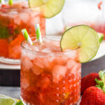 two glasses of Strawberry Caipiroska with crushed ice and two straws garnished with a slice of lime surrounded by fresh strawberries and a lime wedge