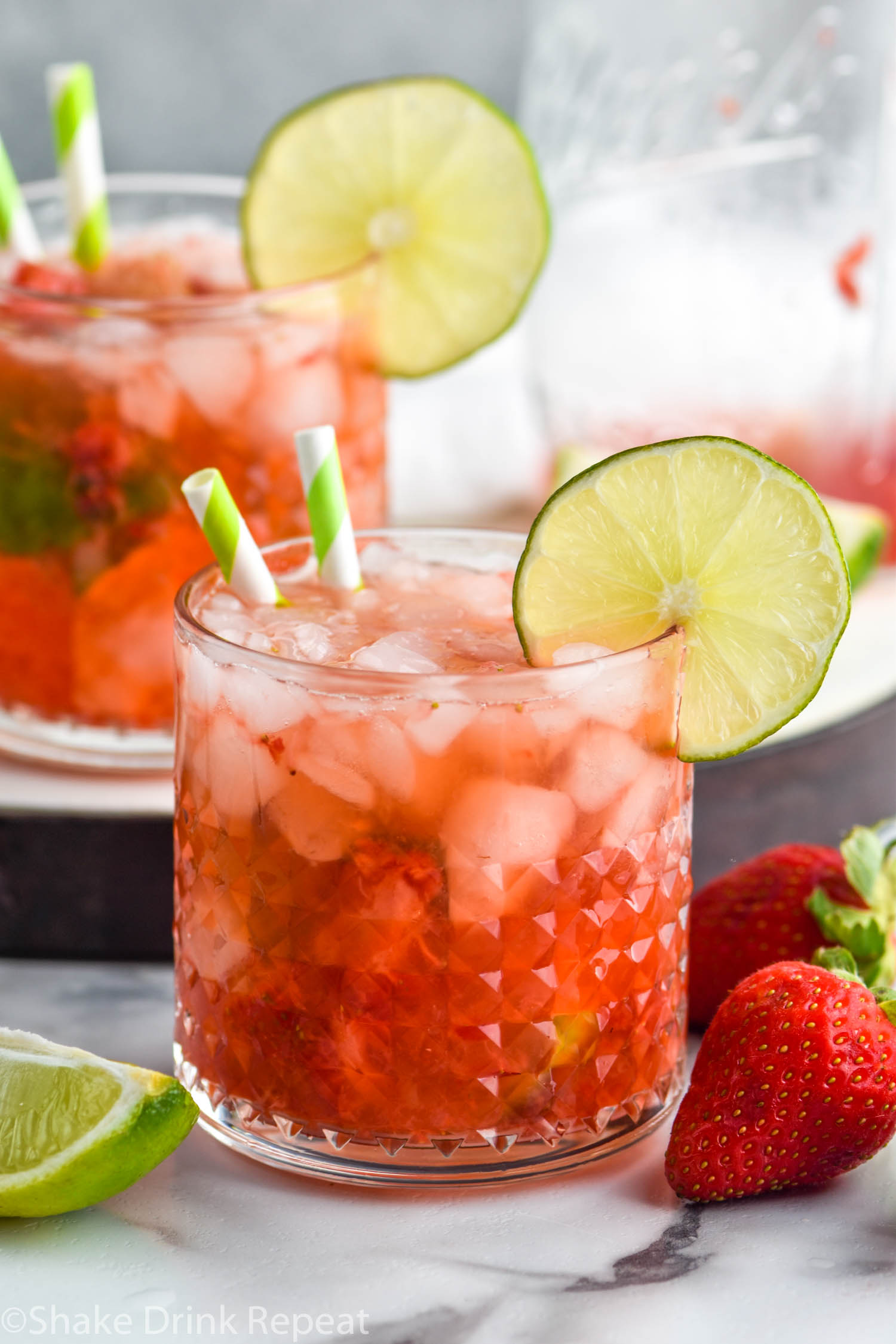 two glasses of Strawberry Caipiroska with crushed ice and two straws garnished with a slice of lime surrounded by fresh strawberries and a lime wedge