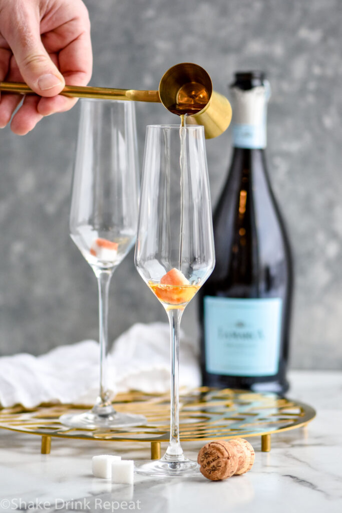 Man's hand pouring a cocktail jigger of Cognac into a champagne flute of bitters and a sugar cube to make a Champagne Cocktail with bottle of champagne in the background