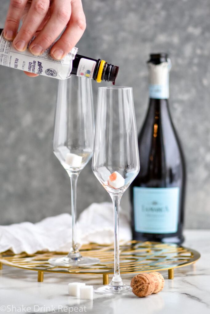 man's hand pouring bottle of cocktail bitters into a champagne flute with a sugar cube to make a Champagne Cocktail recipe with bottle of champagne in the background