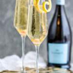 two champagne flutes of Champagne Cocktail garnished with a lemon twist with bottle of champagne in the background