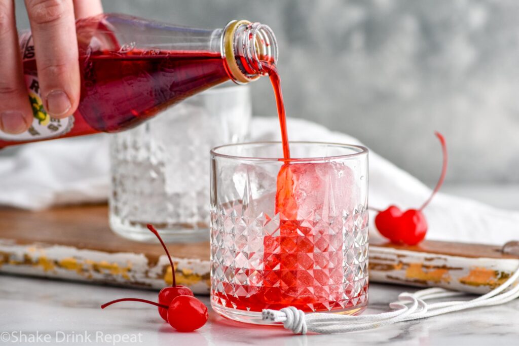 man's hand pouring a bottle of grenadine into a glass of ice to make a Dirty Shirley drink recipe surrounded by maraschino cherries