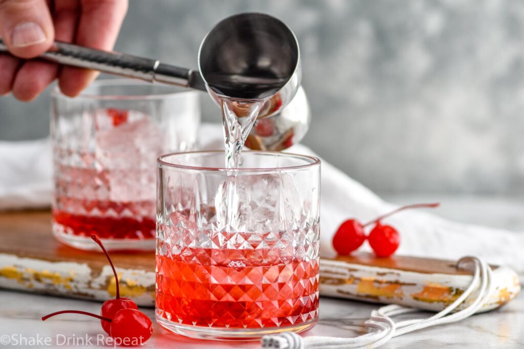 man's hand pouring cocktail jigger of vodka into a glass of Dirty Shirley ingredients surrounded by maraschino cherries