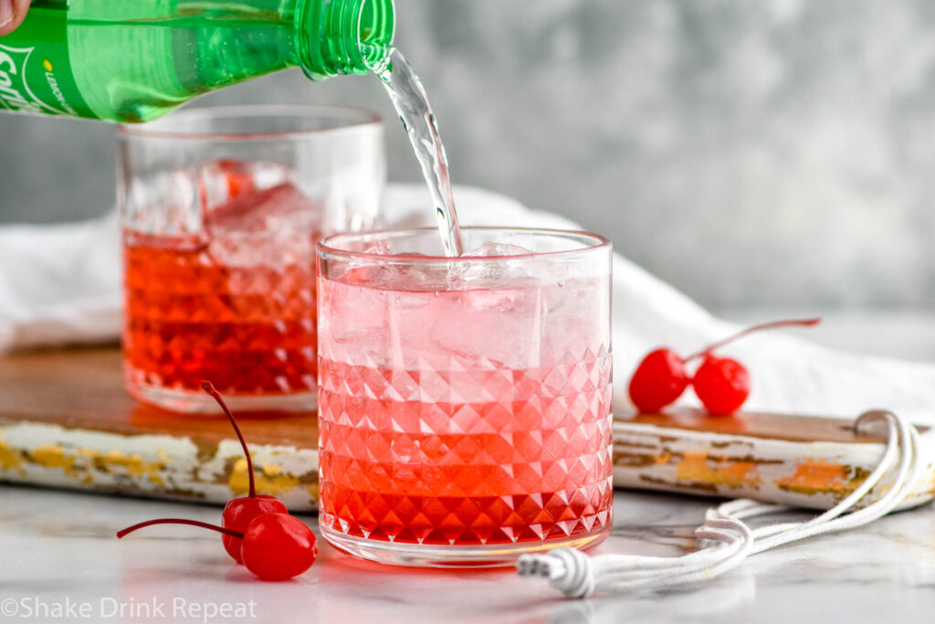 bottle of Sprite pouring into a glass of Dirty Shirley ingredients surrounded by maraschino cherries