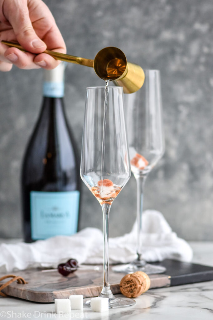 man's hand pouring cocktail jigger of Gin into a champagne flute to make a Gin Champagne Cocktail with bottle of champagne in the background