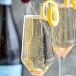 two champagne flutes of Gin Champagne Cocktail garnished with a lemon twist and cherry with bottle of champagne in the background