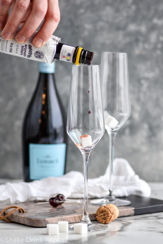 Man's hand pouring bottle of bitters into a champagne flute with sugar cube to make a Gin Champagne Cocktail recipe with bottle of Champagne in the background