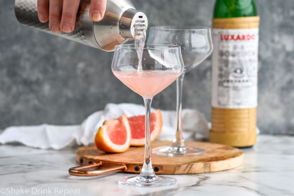 Man's hand pouring cocktail shaker of Hemingway Daiquiri ingredients pouring into a glass, garnished with a grapefruit peel twist, with a bottle of Luxardo in the background