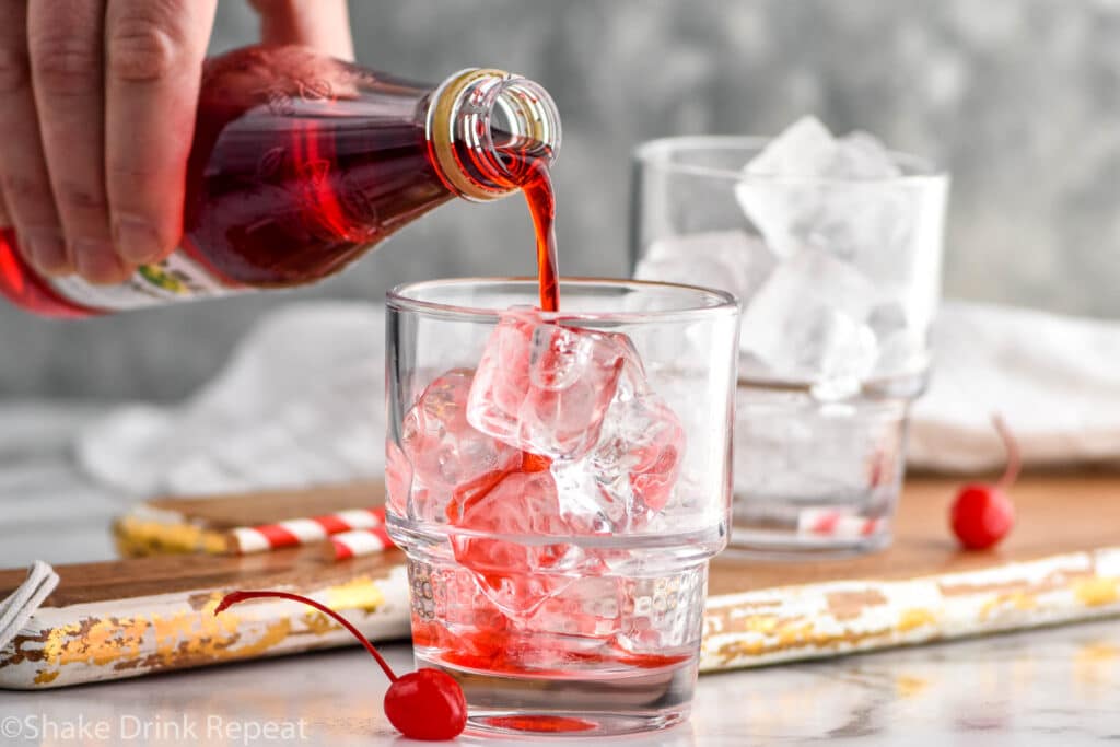 Man's hand pouring bottle of grenadine into a glass of ice to make a Shirley Temple drink with maraschino cherries in front of glass