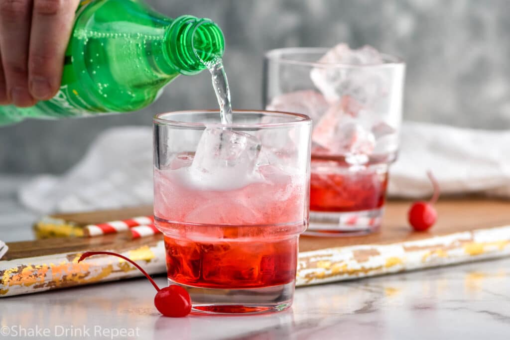 man's hand pouring bottle of Sprite into a glass of Shirley Temple ingredients and ice surrounded by maraschino cherries