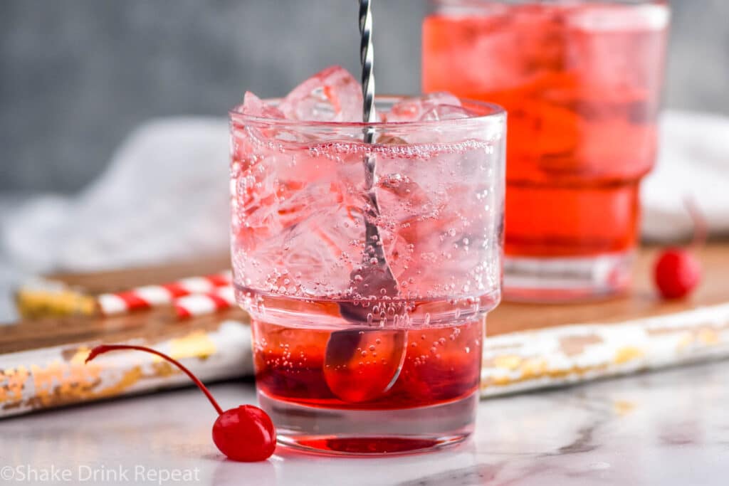 close up of stirring a glass of shirley temple recipe with ice surrounded by a maraschino cherry