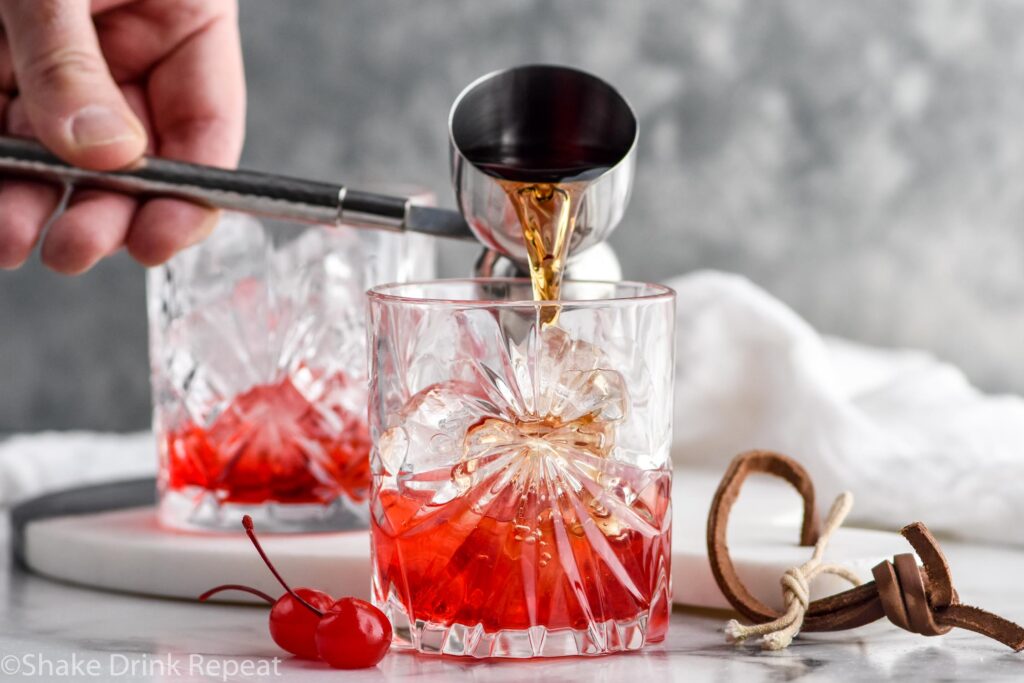 man's hand pouring jigger of rum into a glass of ice and grenadine to make a Shirley Temple Black drink with two maraschino cherries