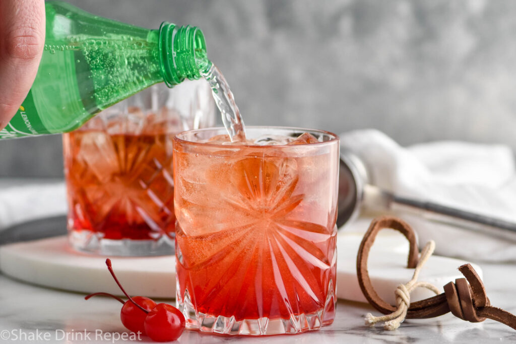 man's hand pouring a bottle of Sprite into a glass of Shirley Temple Black with ice surrounded by maraschino cherries