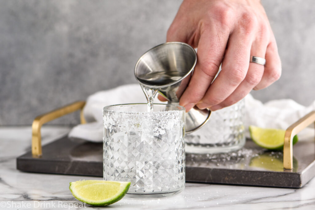 man's hand pouring jigger of tequila into a glass of ice with salted rim to make a tequila and ginger ale cocktail