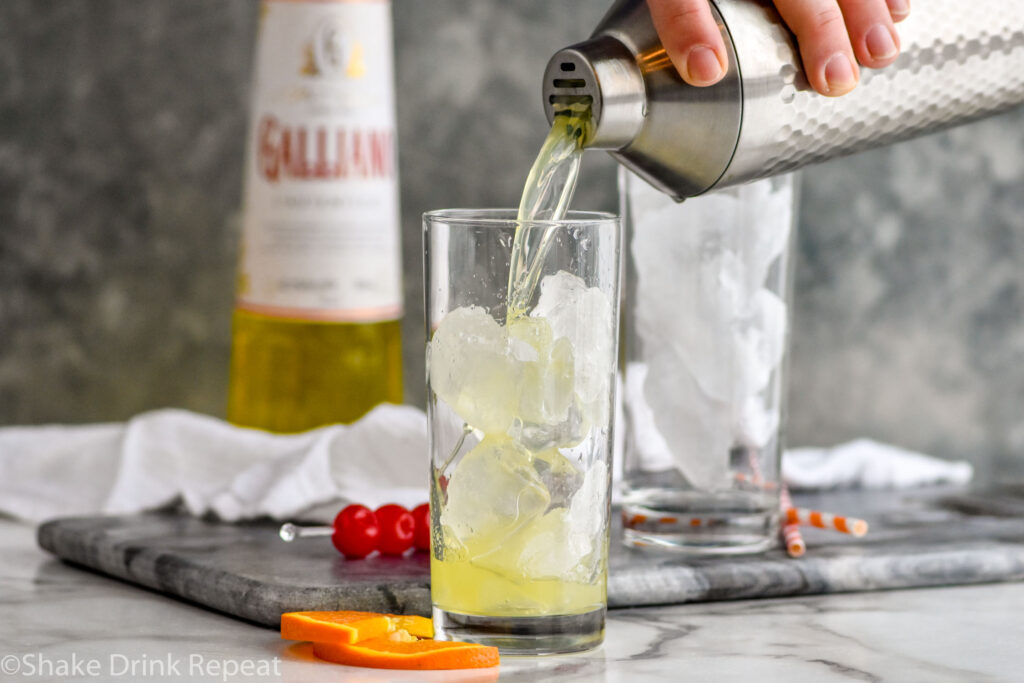 man's hand pouring cocktail shaker of Yellow Bird Drink ingredients into a glass of ice with bottle of Galliano in the background