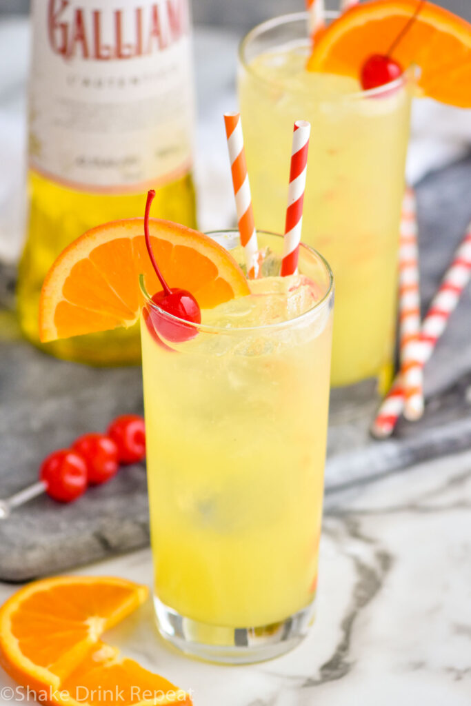 Two glasses of Yellow Bird Drink recipe with ice and two straws, garnished with a cherry and orange slice with bottle of Galliano in the background