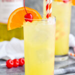 Two glasses of Yellow Bird Drink recipe with ice and two straws, garnished with a cherry and orange slice with bottle of Galliano in the background