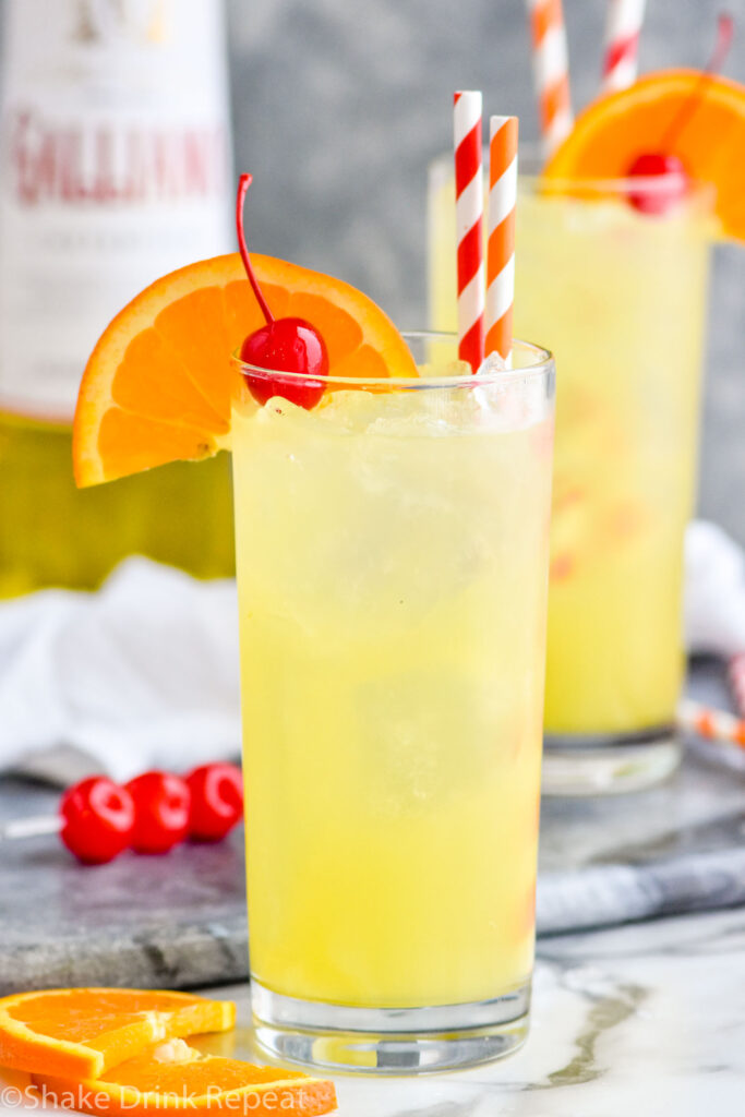 Two glasses of Yellow Bird Drink recipe with ice and two straws, garnished with a cherry and orange slice with bottle of Galliano in the background