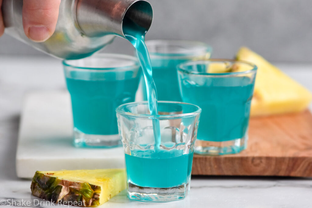 cocktail shaker of Blue Scooby Snack Shot ingredients pouring into a shot glass surrounded by fresh pineapple