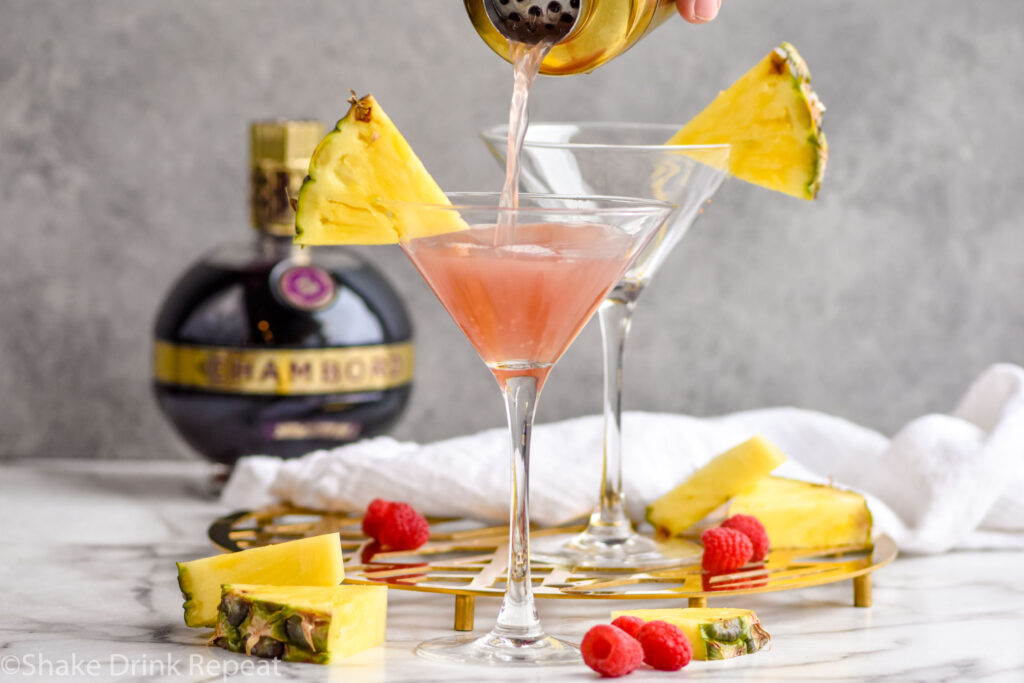 cocktail shaker pouring French Martini ingredients into a martini glass with fresh pineapple, surrounded by fresh raspberries, pineapple, and a bottle of raspberry liqueur.