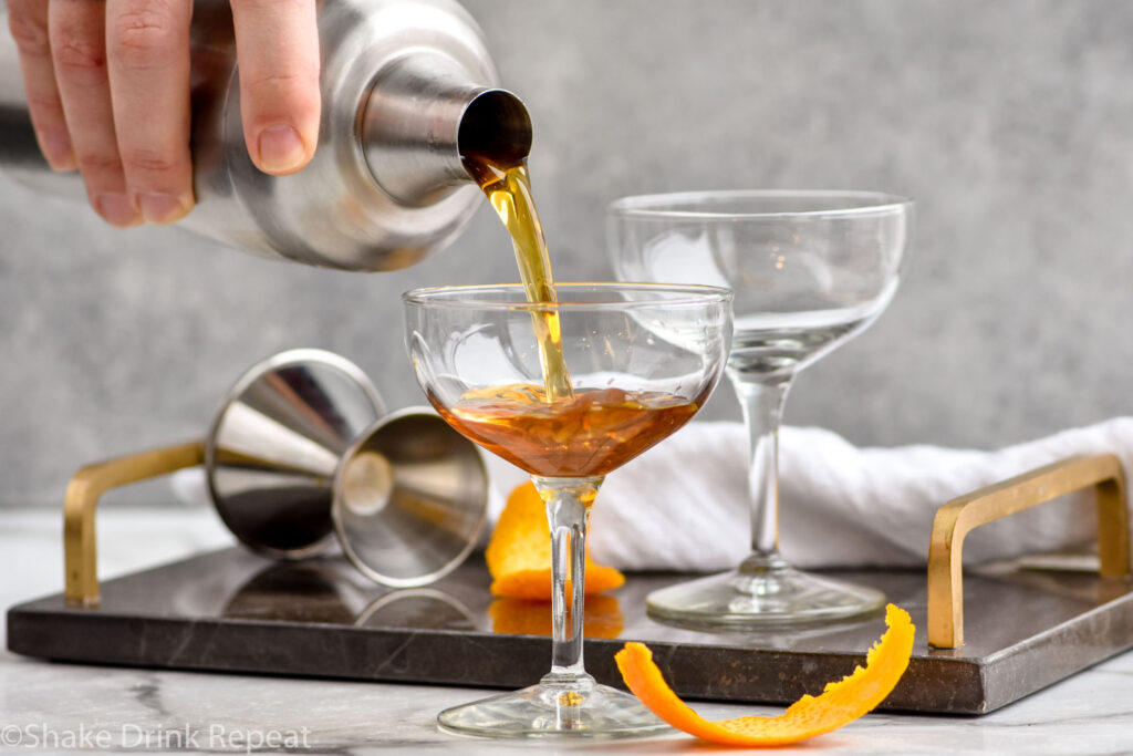 man's hand pouring cocktail shaker of Hanky Panky ingredients into a coupe glass next to a orange peel garnish
