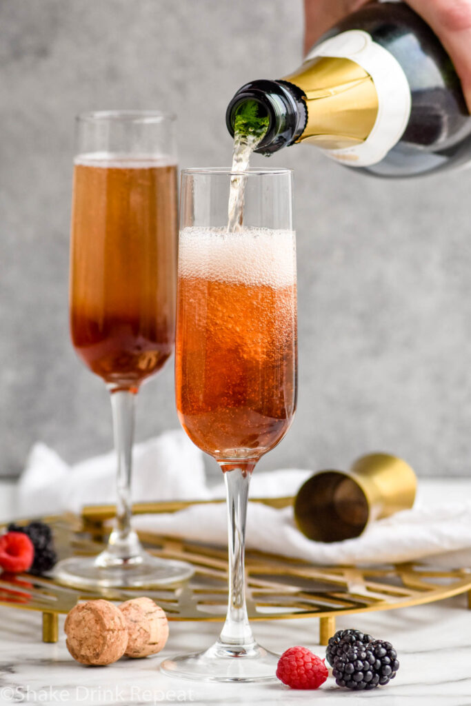 man's hand pouring bottle of champagne into a champagne flute of Crème de cassis to make a Kir Royale cocktail surrounded by fresh raspberries and blackberries