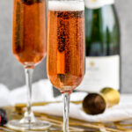 two champagne flutes of Kir Royale cocktail surrounded by fresh raspberries and blackberries and bottle of champagne