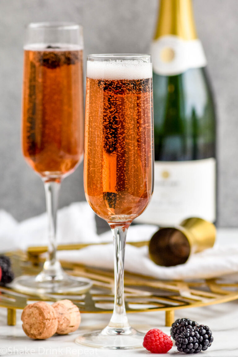 two champagne flutes of Kir Royale cocktail surrounded by fresh raspberries and blackberries and bottle of champagne