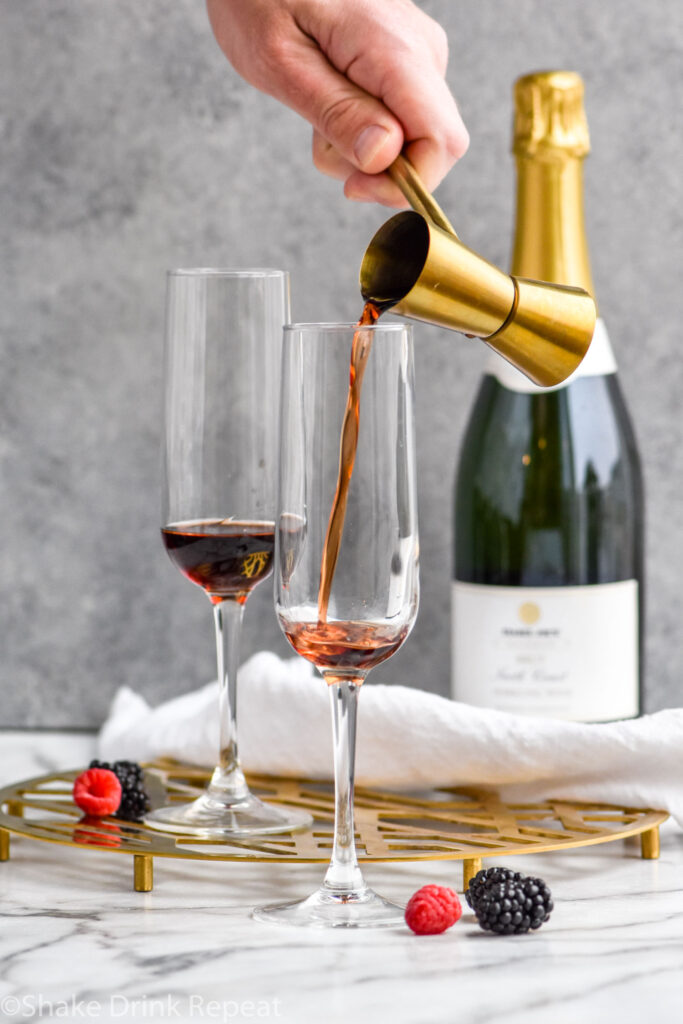 man's hand pouring cocktail jigger of Crème de cassis into a champagne flute to make a Kir Royale recipe with bottle of champagne in the background