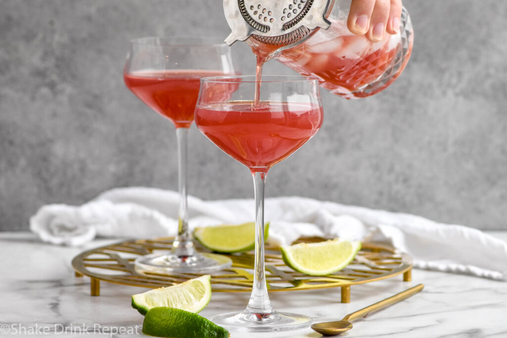 man's hand pouring mixing glass of Scarlett O'Hara Cocktail ingredients with ice into a glass surrounded by lime wedges