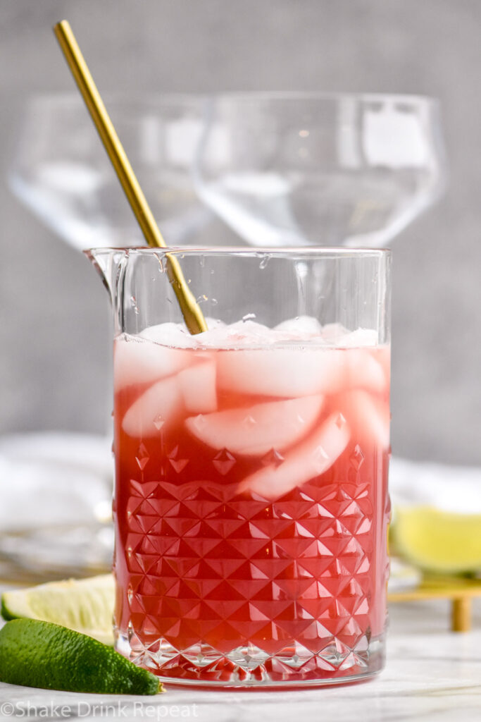 Mixing glass of Scarlett O'Hara Cocktail with ice surrounded by lime wedges and two empty glasses