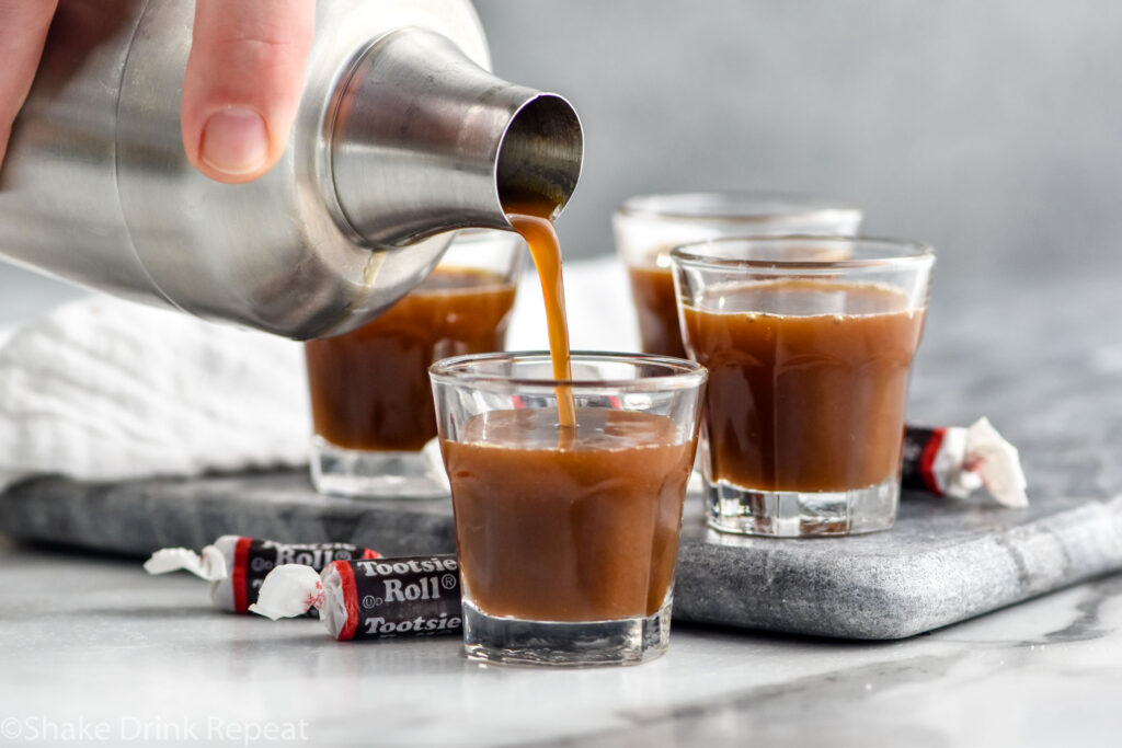 man's hand pouring cocktail shaker of Tootsie Roll Shot ingredients into a shot glass surrounded by Tootsie Roll candies