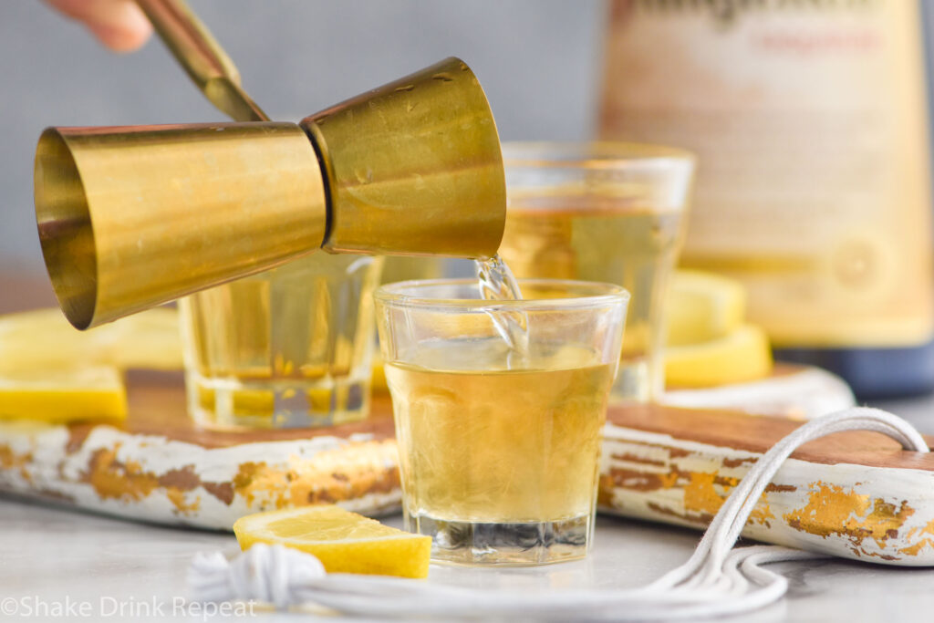 man's hand pouring cocktail jigger of hazelnut liqueur into a shot glass to make a chocolate cake shot recipe surrounded by lemons