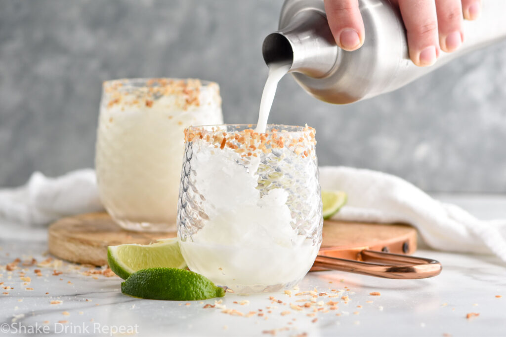 man's hand pouring cocktail shaker of Coconut Margarita ingredients into a glass of ice and rimmed with toasted coconut surrounded by lime wedges