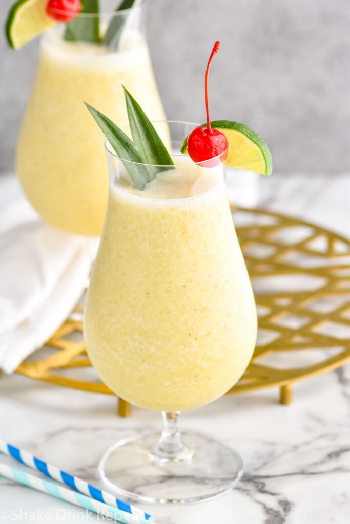 two hurricane glasses of frozen Banana Daiquiri garnished with a cherry, lime, and leaves