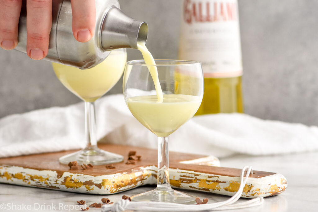 man's hand pouring cocktail shaker of Golden Cadillac recipe into a glass with bottle of Galliano in the background