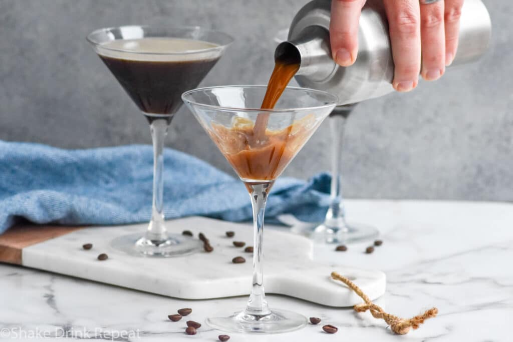 man's hand pouring cocktail shaker of Espresso Martini ingredients into a martini glass surrounded by espresso beans