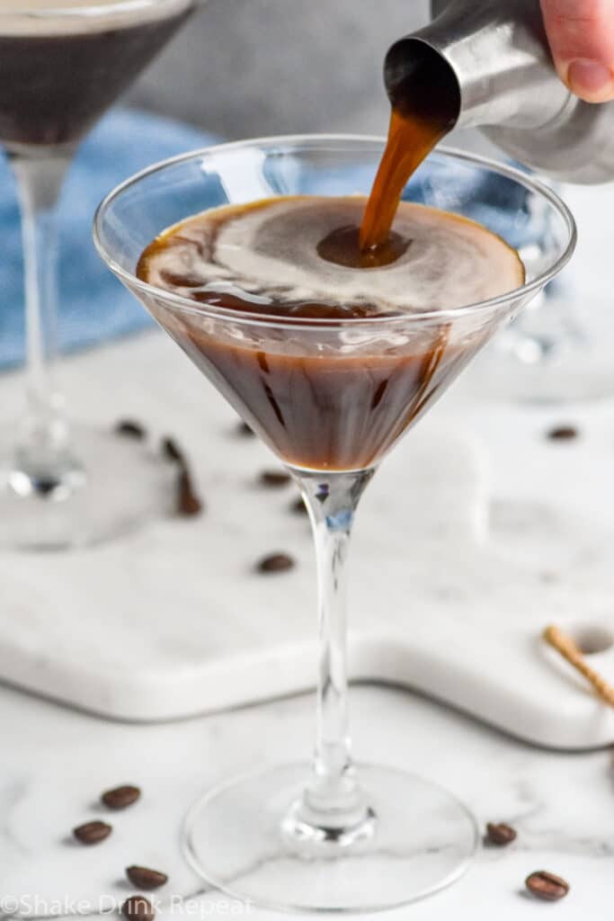 cocktail shaker pouring Espresso Martini ingredients into a martini glass surrounded by espresso beans
