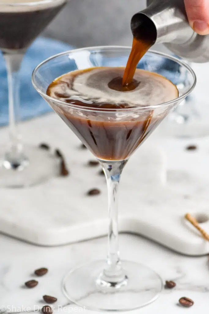 cocktail shaker pouring Espresso Martini ingredients into a martini glass surrounded by espresso beans